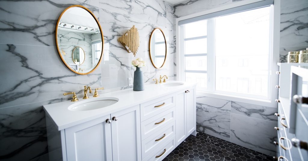 White and marble bathroom with white shaker vanity and gold framed round mirrors.