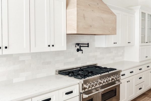 White kitchen with gray and white subway tile backsplash, light wood range hood, and black hardware.